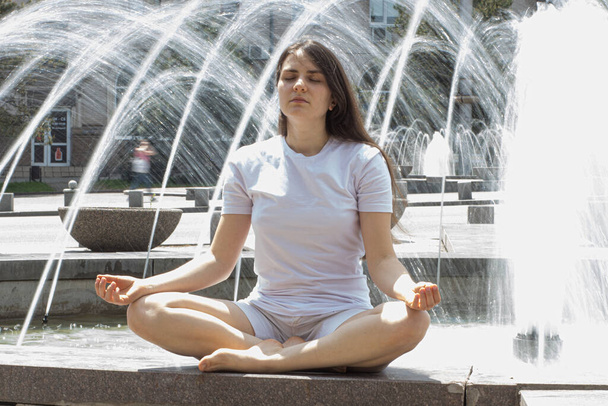 Красивая брюнетка в белой одежде медитирует возле фонтана на улице в городе. Йога и медитация, замедляющиеся в ритме современной жизни - Фото, изображение