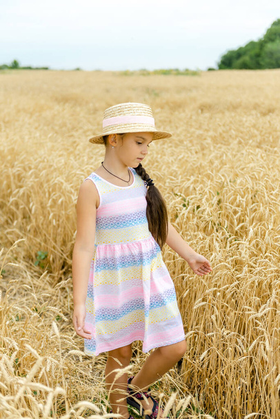 Una niña con un sombrero de paja camina a través de un campo de trigo y acaricia espigas de trigo con sus manos. - Foto, imagen