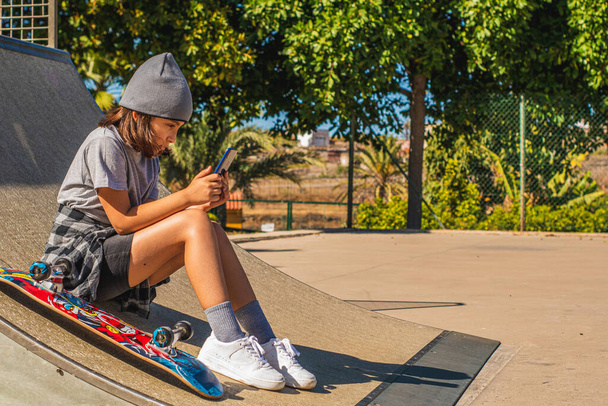 Caucasian girl sitting on a skateboard ramp with her board beside her, while looking at her mobile phone - Foto, Bild