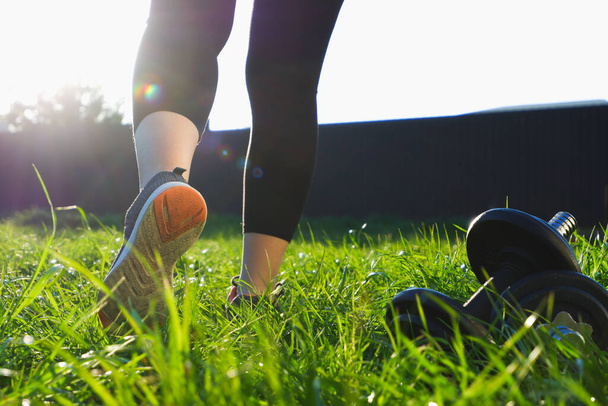 Treino ao ar livre. Uma mulher caminha sobre a grama na luz de fundo do sol poente. Visão traseira das pernas em tênis de corrida close-up. Há halteres por perto.. - Foto, Imagem