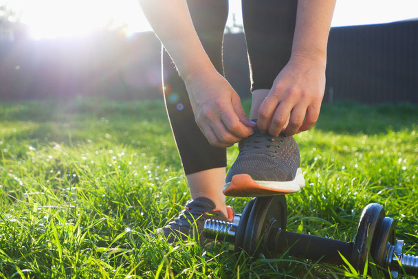 Тренировка на улице. Женщина завязывает шнурки на кроссовках перед пробежкой. Вид на фоне заходящего солнца. Крупный план ног в кроссовках. Колокольчики на зеленой траве. - Фото, изображение