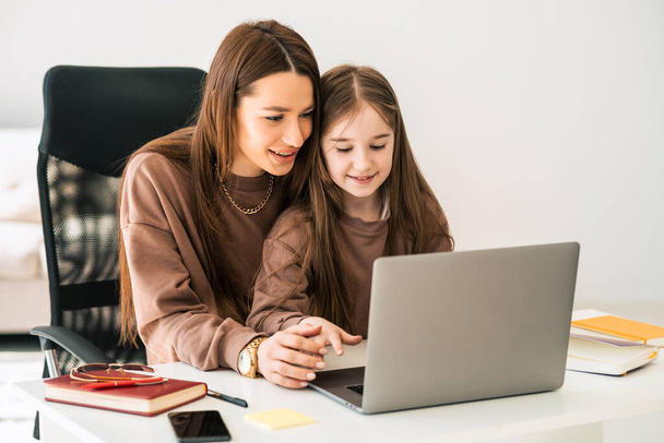 Moeder en dochter doen huiswerk op de laptop. Klein schoolmeisje op afstand thuis leren - Foto, afbeelding