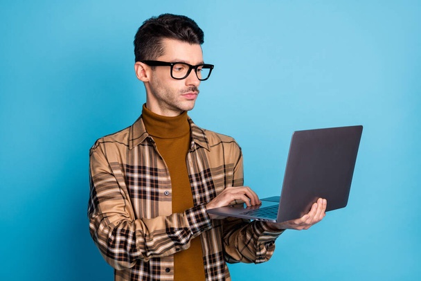 Photo of young serious focused handsome man programmer in glasses working in laptop isolated on blue color background - Fotó, kép