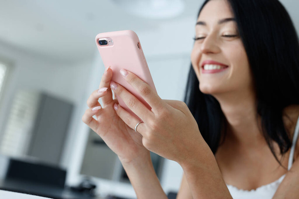 Genç bir kadın, modern pembe akıllı telefonu kullanırken mutlu bir şekilde kendi kendine sırıtıyor. Telefona odaklanmış, yakın eğimli, düşük açılı bir şekilde evde dinleniyor. - Fotoğraf, Görsel