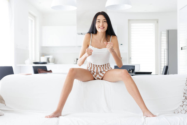 Мотивована збуджена босоніж молода жінка стискає кулаки, коли вона сидить збалансовано на спині дивана у вітальні з високим ключем, дивлячись на камеру з щасливою посмішкою
 - Фото, зображення