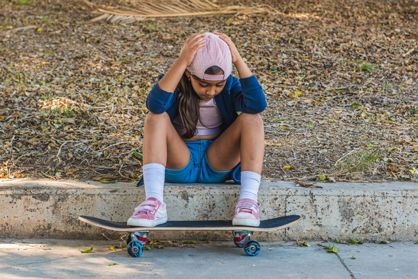 Девушка-фигуристка сидит на ступеньках со своим скейтбордом и кепкой, с грустным или скучным жестом. - Фото, изображение