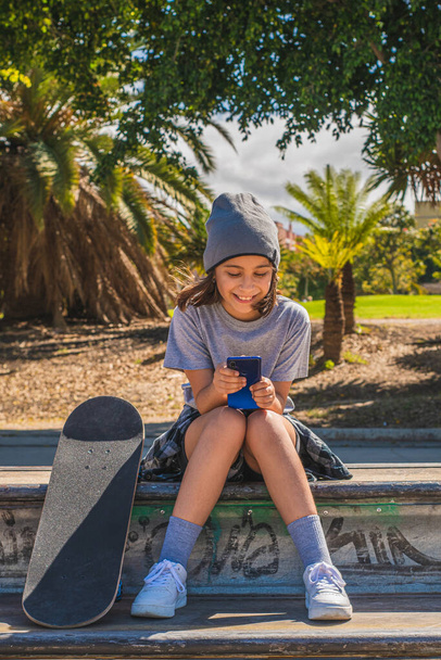 Předpubertální dívka, běloška, sedí na překážce v bruslovém parku, s nohama na hrací desce, zatímco chatuje nebo surfuje na internetu se svým mobilním telefonem, velmi se usmívá. - Fotografie, Obrázek