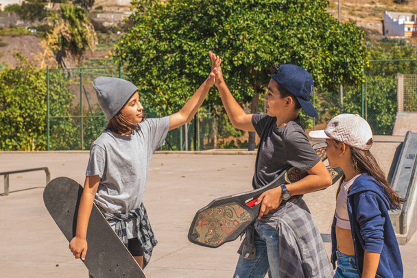 Três crianças de patinação caucasianas, vestidas com roupas casuais e urbanas, cumprimentam-se felizes depois de se encontrarem em um parque de skate. - Foto, Imagem