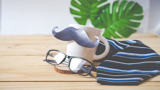 Blur of Background, Happy Father's day concept. Un papel de bigote negro, una corbata azul, una caja de regalo azul y vasos sobre mesa de madera. - Foto, imagen