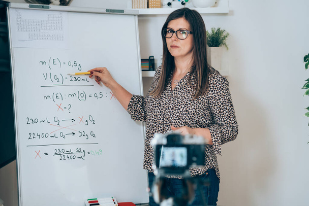 Teacher recording video tutorial for e-learning class.Female professor standing and explaining the assignment pointing on the whiteboard and using camera for live streaming online on the internet for Physic students, remote education class. - Φωτογραφία, εικόνα