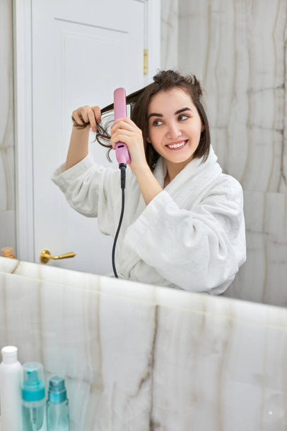 woman using a hair straightener in bathroom. - Fotó, kép