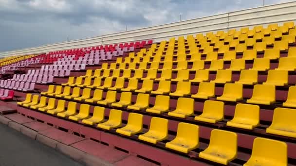 Asientos amarillos, verdes, rojos y azules en fila en el estadio sin el jugador y el público. Asientos vacíos en estadio o pista de carreras durante la pandemia del coronavirus COVID-19. - Metraje, vídeo