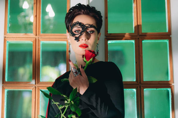 Nahaufnahme einer geheimnisvollen Frau mit schwarzer Spitzenmaske, die sich an eine alte Tür lehnt und eine rote Rose in der Hand hält - Foto, Bild