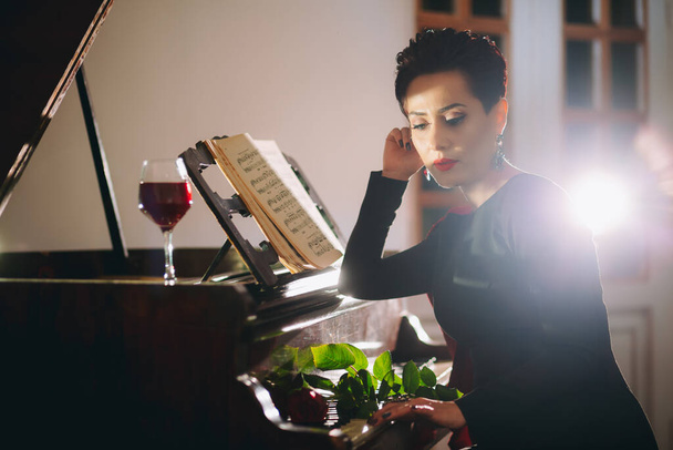 Elegante, romantische en luxe vrouw aan de piano met roos en rood wijnglas. Luxe vrouw poseren aan de piano en creatieve lichtstraal achter - Foto, afbeelding