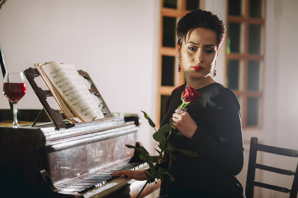 Elegante, romantische und traurige Frau, die mit einer Hand die Klaviertastatur berührt und eine rote Rose in der Hand hält. Kreative Innenbeleuchtung mit schöner Dame - Foto, Bild