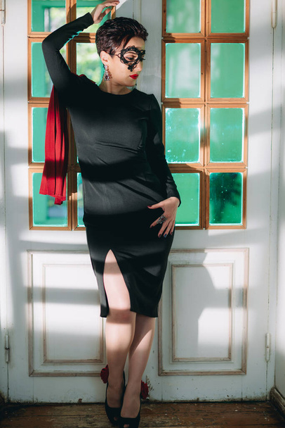 Long shot of stylish, fashionable and mysterious woman with black dress and black lace mask standing in front of vintage door and posing for camera - 写真・画像