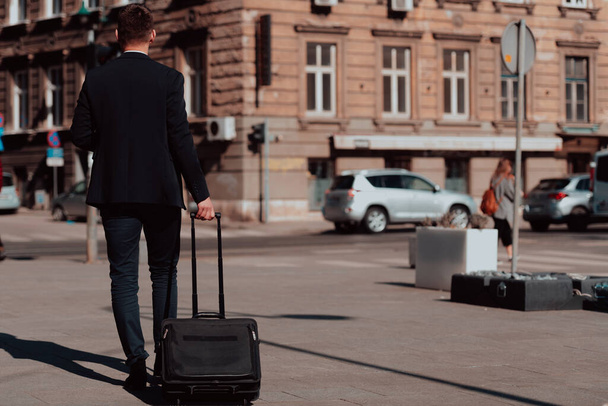 Going to airport terminal. Confident businessman traveler walking on city streets and pulling his suitcase drinking coffee and speaking on smartphone - Foto, Bild