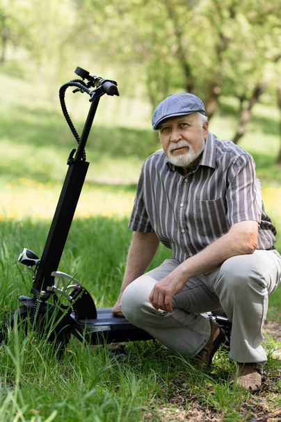 A positive, elderly man went for a walk to the park, stopped and sat down to rest next to an electric scooter. Summer, sunny day. Healthy lifestyle of the elderly. - Photo, Image
