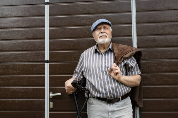 Um homem idoso positivo fica perto da porta e segura uma scooter ecológica ao volante. Estilo de vida saudável dos idosos. - Foto, Imagem