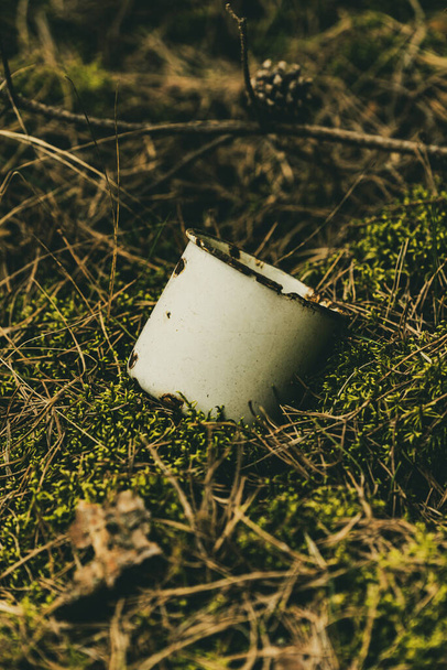 caneca de metal branco com uma aparência ligeiramente enferrujada encontra-se em um musgo macio abandonado em uma floresta de pinheiros - Foto, Imagem
