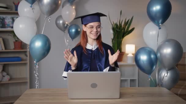 online graduation, young woman in an academic cap and gown rejoices at presentation of diploma in virtual ceremony via video link on laptop while sitting at home background of balloons - Footage, Video