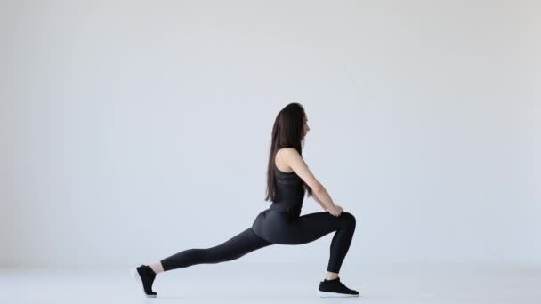 The young athletic brunette in the black gym suit is doing the single leg lunges and raising hands up at the white background. - Footage, Video