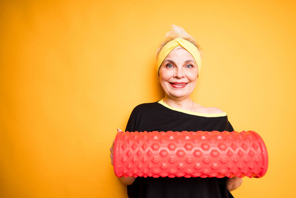 Cheerful elderly woman holding fascia in hands while getting ready for training and standing on yellow background - Fotó, kép