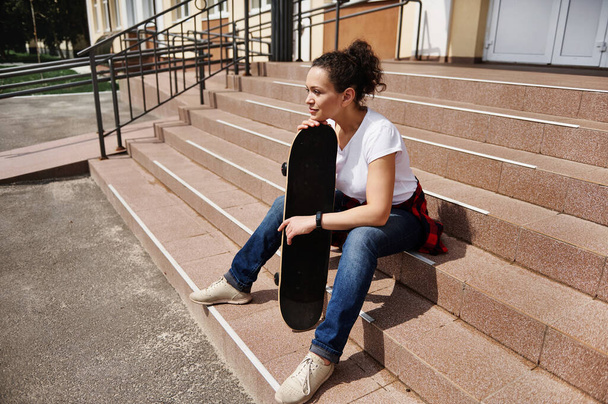 Portrait of beautiful young african american woman with skateboard sitting on steps of unrecognizable building - Zdjęcie, obraz