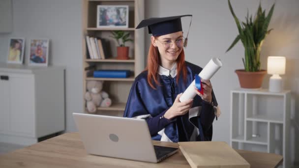 Выпускница с дипломом в руках радуется на онлайн церемонии окончания университета, используя веб-камеру на ноутбуке, сидя дома на фоне воздушного шара - Кадры, видео