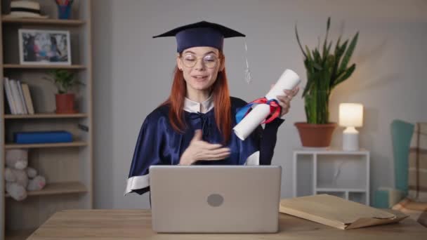 opiskelu verkossa, nuori nainen akateemisissa vaatteissa iloitsee tutkintotodistuksesta, jonka hän sai etäopetuksen aikana istuessaan kannettavassa huoneessa - Materiaali, video