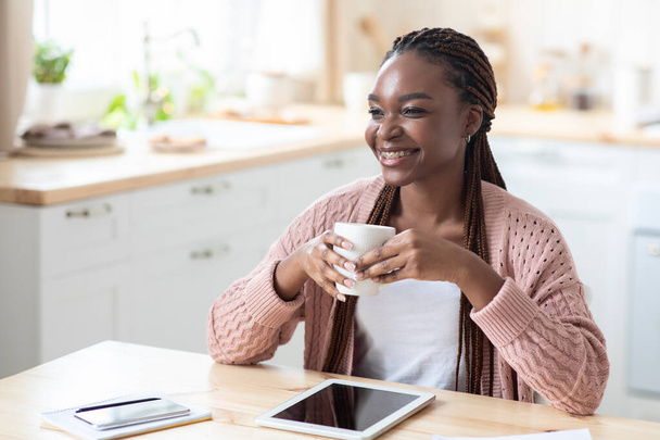 Afro-Amerikan kadın kahve içiyor ve mutfakta dijital tablet kullanıyor. - Fotoğraf, Görsel