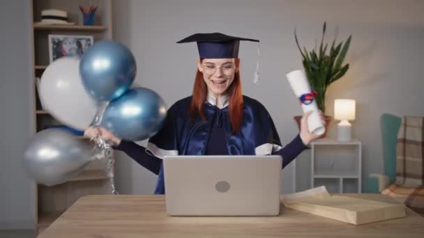 graduación en línea, emocional chica feliz estudiante universitario en un vestido académico se regocija por el diploma recibido y sopla certificado como la melodía y ondear globos mientras está sentado en el ordenador portátil en casa - Imágenes, Vídeo