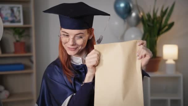 online training, portrait of joyful female graduate in an academic gown and hat talking by video call on laptop and showing diploma - Video, Çekim