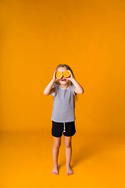 小さなブロンドの女の子が立って目を閉じてオレンジの半分テキストのためのスペースを持つ黄色の背景に。夏の子 - 写真・画像