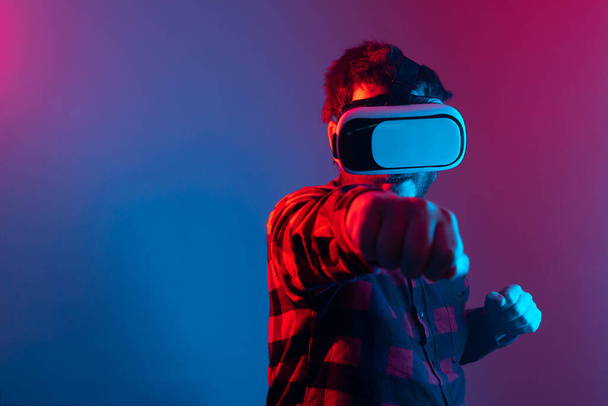 VRメガネをかけた青年が空中でパンチを放り投げ。仮想現実のビデオゲーム。ネオンとスタジオの肖像画. - 写真・画像