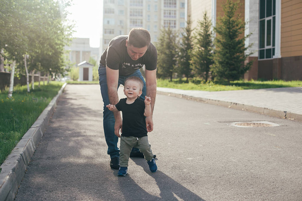 父と息子が一緒に歩いている。赤ちゃんは父親の助けを借りて最初の一歩を踏み出す。本物の瞬間、夏のライフスタイル - 写真・画像