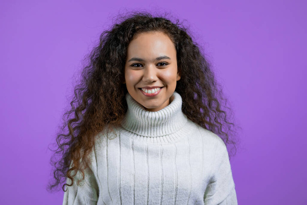 Portrait of pretty woman on violet studio background. Positive young girl with natural curly hair, she in white sweater smiles to camera. - Фото, изображение