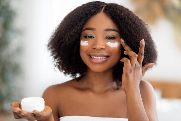 自宅でミラーの近くの瓶から保湿顔クリームを適用する官能的な若い黒人女性。美容・天然化粧品 - 写真・画像