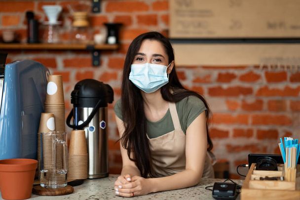 Femme arabe amicale dans le tablier debout près du comptoir du bar et attendant les clients, portant un masque de protection - Photo, image