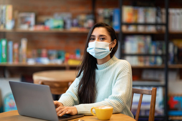 Young arab woman in mask searching job during covid-19 pandemic, using laptop in cafe, checking vacancies in internet - Fotoğraf, Görsel