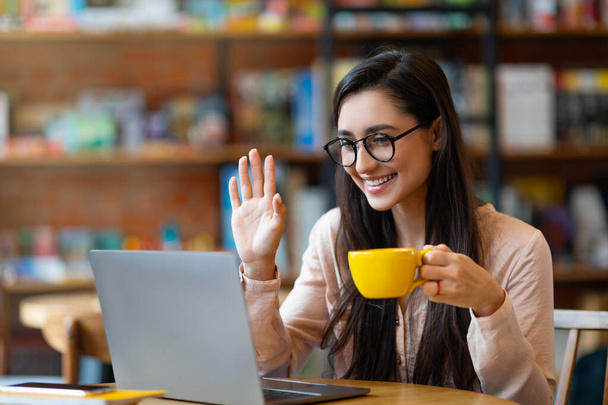 Vriendelijke latijnse vrouw met videogesprek van cafe, zwaaiende hand naar laptop camera en het drinken van warme drank - Foto, afbeelding