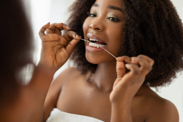 Jolie jeune femme noire se nettoie les dents avec du fil dentaire devant le miroir à la maison. Hygiène buccale et bien-être - Photo, image