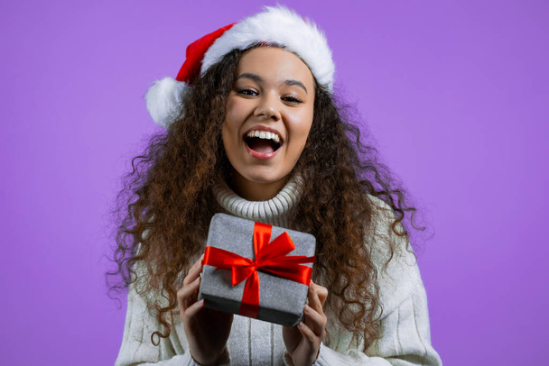 Beautiful woman in Santa hat received gift box with bow. She is happy and flattered by attention. Girl in sweater smiling with present on purple background. New year, Christmas concept. - 写真・画像