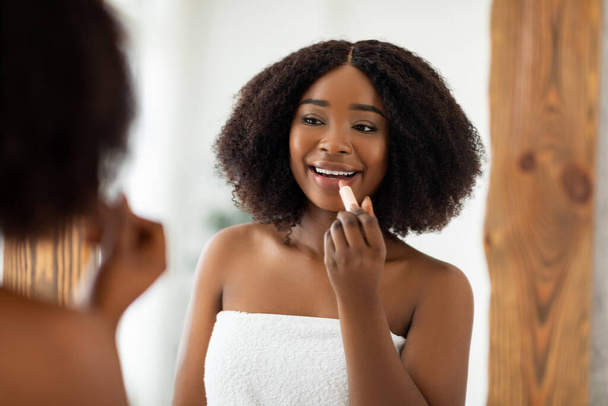 Beauty portrait of pretty young black woman applying lipstick and looking at mirror indoors. Organic makeup products - Photo, Image