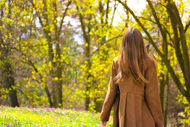 Μια νεαρή γυναίκα απολαμβάνει μια βόλτα σε ένα ανθισμένο λιβάδι σε ένα ανοιξιάτικο πάρκο. Πίσω όψη μιας κυρίας με όμορφα καστανά μαλλιά σε ένα καφέ παλτό στη φύση. Άνθρωποι έξω. Έννοια ηρεμίας και χαλάρωσης. - Φωτογραφία, εικόνα