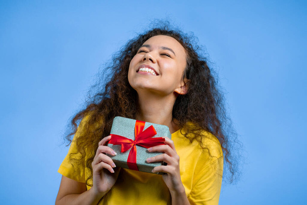 Beautiful woman received gift box with bow. She is happy and flattered by attention. Girl smiling with present on blue background. Studio portrait - Foto, imagen