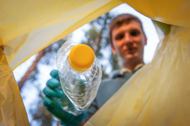 Raccolta di rifiuti in natura. Giovane uomo mette spazzatura di plastica in una borsa gialla, vista interna - Foto, immagini