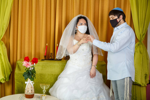 During a chuppah ceremony at a Jewish wedding in a synagogue, the groom puts a ring on the bride's index finger of a newlywed couple wearing a pandemic mask. Horizontal photo - Fotó, kép