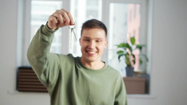 Een jongeman staat in een witte lege kamer en schudt sleutels met een glimlach - Foto, afbeelding