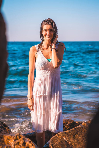 A young Caucasian girl smiling in a white dress and wet hair sitting on a rock by the sea, summer lifestyle, vertical photo - Fotografie, Obrázek